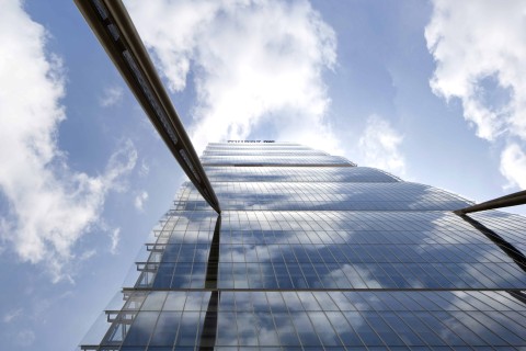 Allianz Tower aus der Froschperspektive. Der Himmel ist klar mit erinnerten Wolken. Die Fassade des Hochhauses ist aus Glas, an ihr ragen zwei Stahlträger.