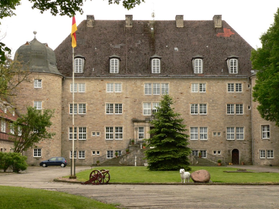 Schloss in bräunlichen Farben vor einem Innenhof mit Rasenflächen und Baum in der Mitte.____