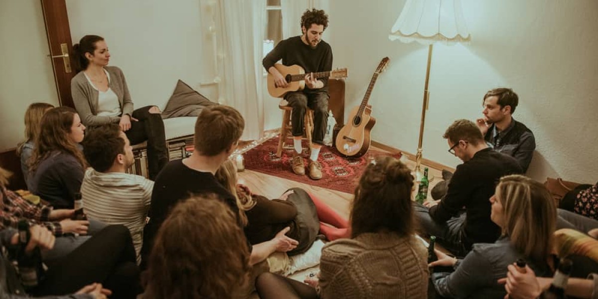 Junge Menschen in einem Wohnzimmer. Ein bärtiger junger Mann spielt gerade Gitarre, die anderen hören gespannt zu.
